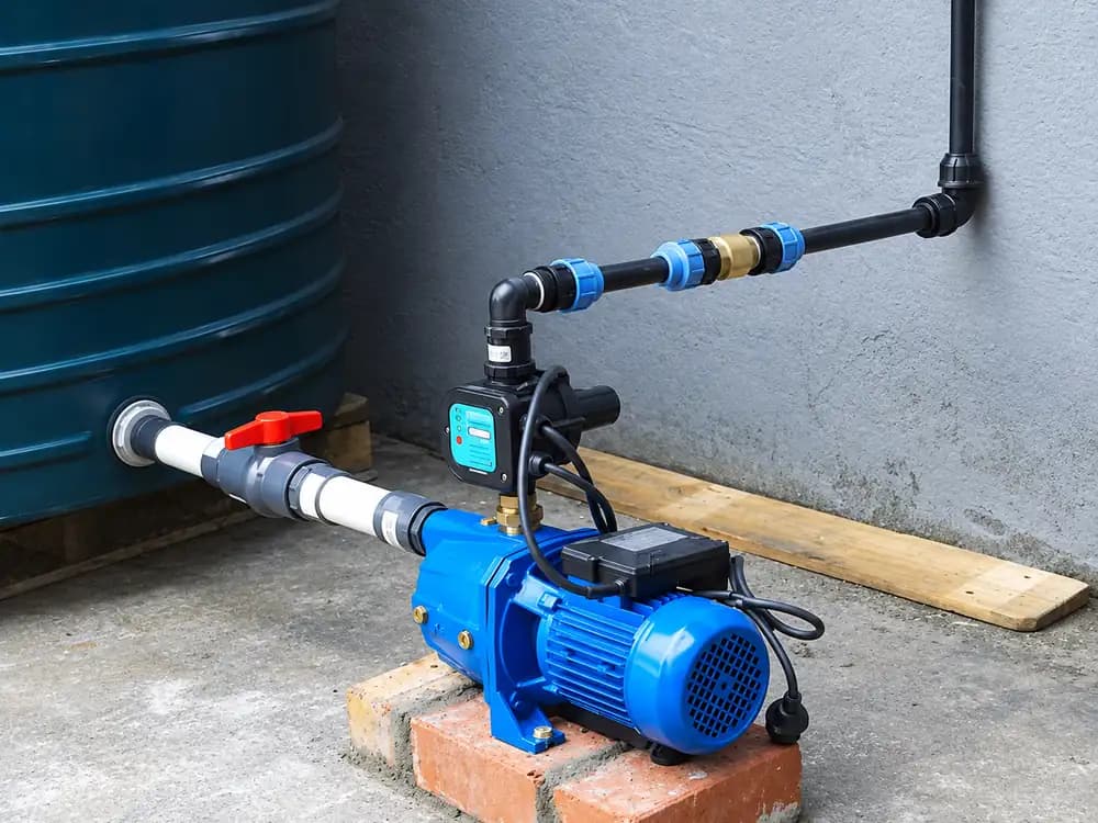 Bomba presurizadora azul conectada a un tanque de agua con válvula y tubería, sobre una base de ladrillo