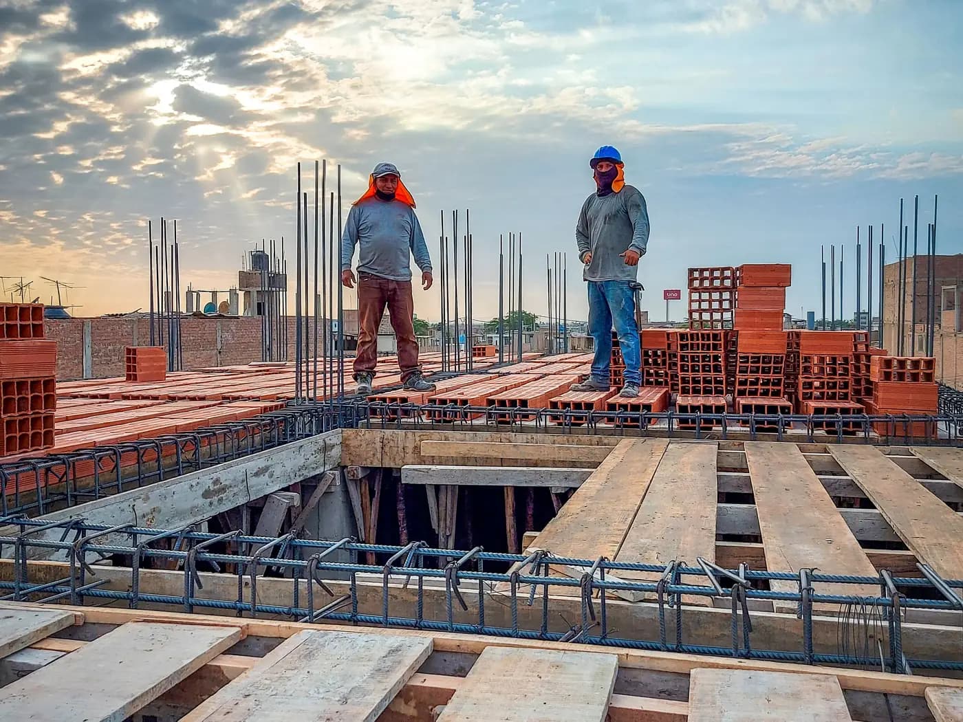 Dos obreros sobre el encofrado de una losa, con acero de refuerzo y ladrillo apilado en una obra de construcción